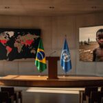 Sala de conferência em Brasília com bandeiras do Brasil e ONU, representando críticas a guerras, fome global e descrédito da ONU.