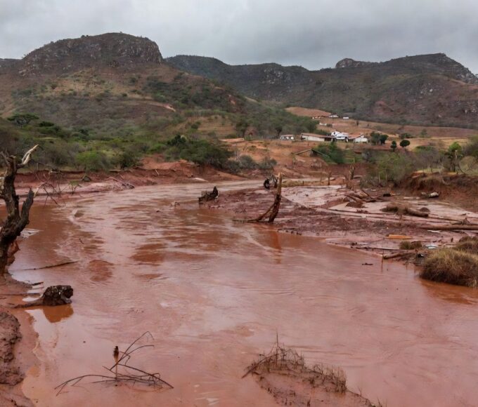 Paisagem devastada por tragédia ambiental em Minas Gerais, com rio poluído e vegetação destruída, representando vítimas que recebem auxílio.