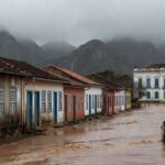 Rua inundada em cidade de Minas Gerais após enchentes, com bandeira brasileira simbolizando auxílio de R$ 7.300 para vítimas.