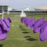 Evento esportivo contra feminicídio no DF, com bandeiras roxas e arquitetura de Brasília em estilo fotojornalístico.