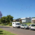 Viaturas da PMDF em rua de Brasília com drones caindo sobre residência, em estilo fotojornalístico.